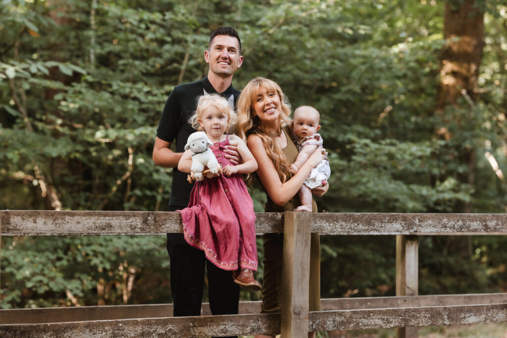 A smiling family of four stands on a wooden fence in a wooded area. The father stands behind the mother, who holds a baby. A young girl in a pink dress holds a stuffed animal and stands next to them. Trees fill the background.