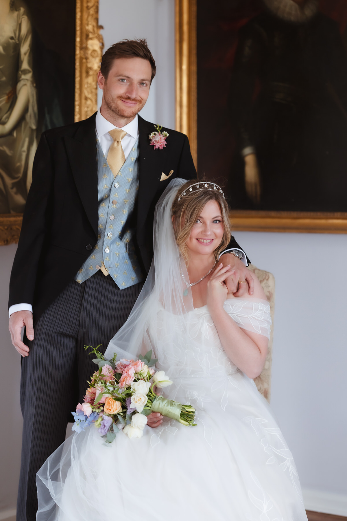 A bride in a white dress and veil sits holding a pastel bouquet while a groom in a suit with a light blue vest stands beside her, smiling, in front of large classical paintings.
