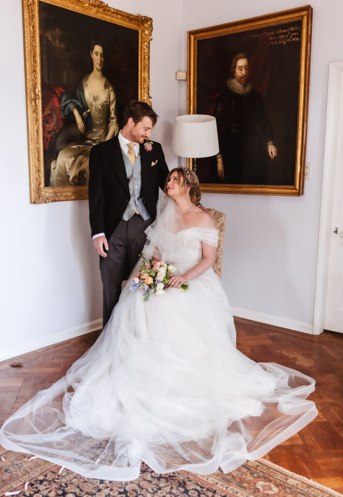 A bride in a white gown sits with a bouquet while a groom in a formal suit stands beside her, gazing at her. They are indoors near two large, framed vintage portraits on the wall.