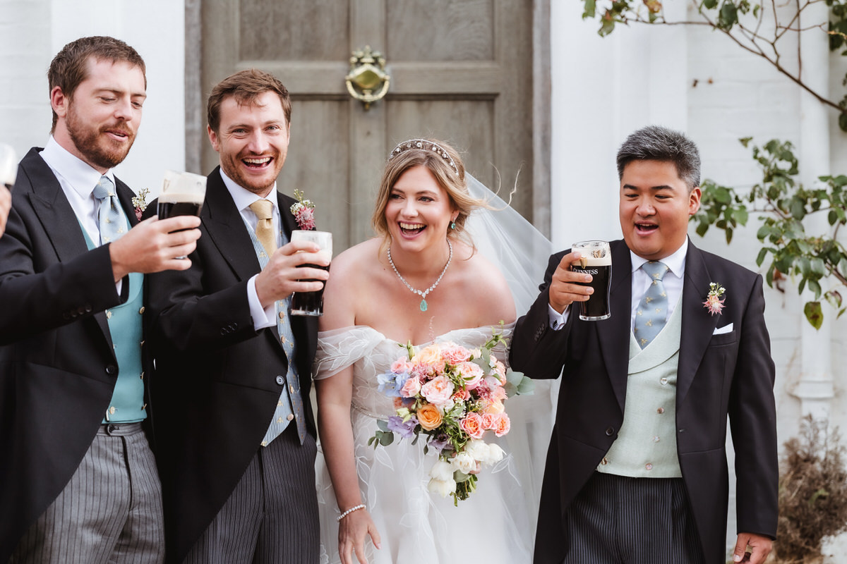 A bride in a white dress and three groomsmen in suits smile and raise pints of dark beer together outdoors, celebrating in front of a wooden door decorated with greenery.