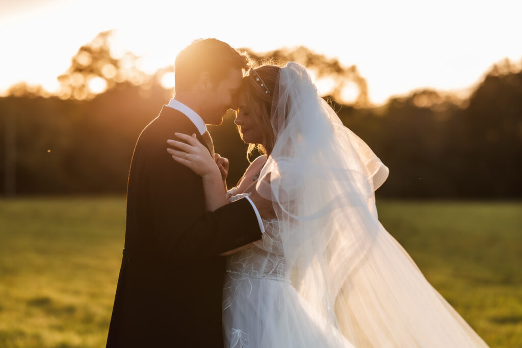 A bride and groom stand close together outdoors at sunset, touching foreheads and embracing, with the bride’s veil flowing and sunlight glowing behind them in a green field.
