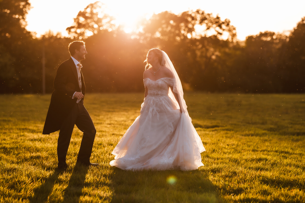 A bride in a white dress and veil stands on sunlit grass, smiling at a groom in a suit. The sun sets behind them, casting a warm golden glow over the scene with trees in the background.