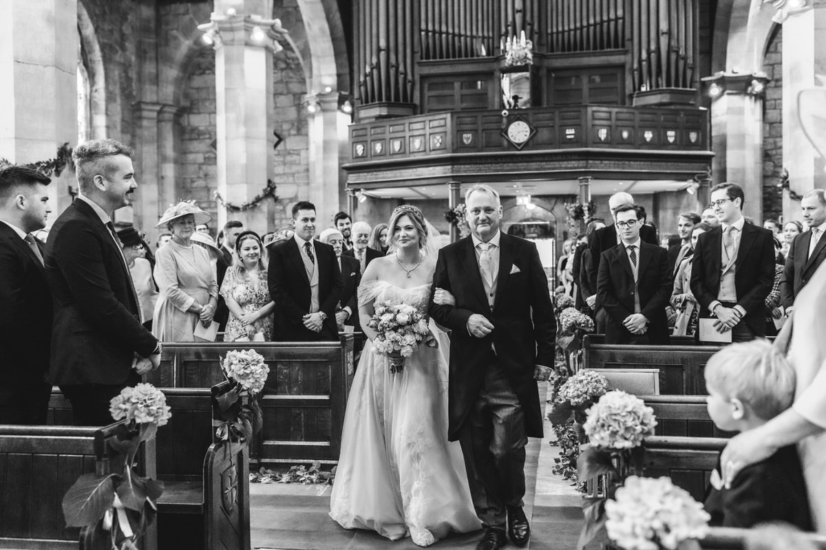 A bride in a white dress, holding a bouquet, walks down the aisle with an older man in a suit inside a church filled with seated and standing guests, who are smiling and watching. The scene is in black and white.