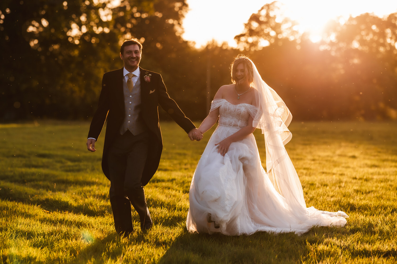 A bride and groom walk hand in hand across a grassy field at sunset, smiling. The bride wears a white gown and veil, and the groom is in a dark suit with a vest and tie. Warm sunlight glows in the background.