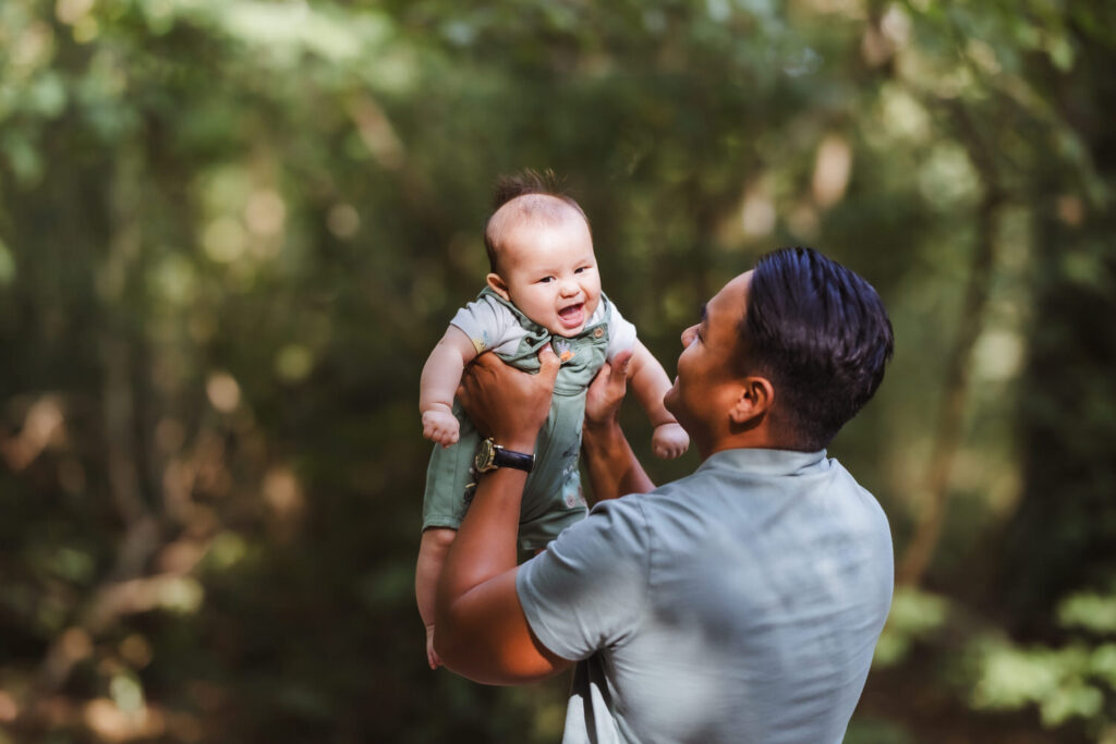 A man in a light green shirt holds a smiling baby up in the air outdoors, surrounded by lush greenery and soft sunlight.