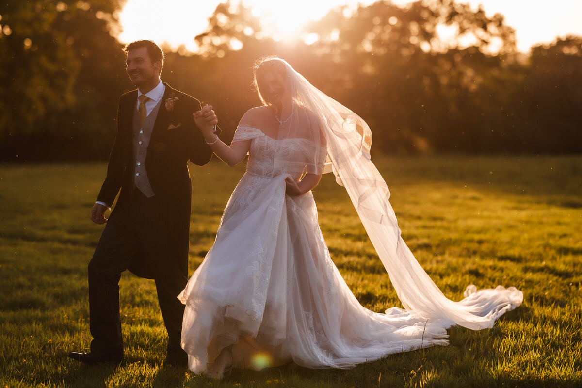 A bride and groom walk hand in hand through a sunlit grassy field at sunset. The bride’s veil flows behind her, and both are smiling, dressed in formal wedding attire. Trees are visible in the background.