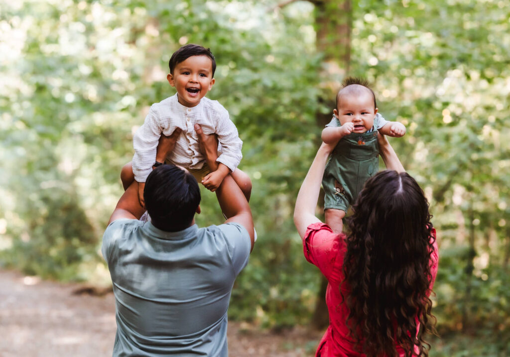 Two adults, seen from behind, lift two smiling young children in the air while standing on a forest path. The background is filled with lush green trees and sunlight filters through the leaves.