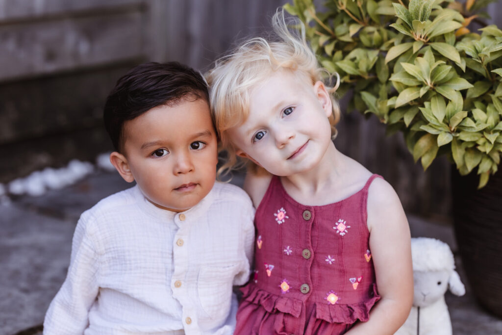 Two young children, one with dark hair and tan skin wearing a light shirt, and one with blonde hair and fair skin wearing a pink dress, sit close together outdoors, smiling softly. A stuffed toy lamb is beside them.