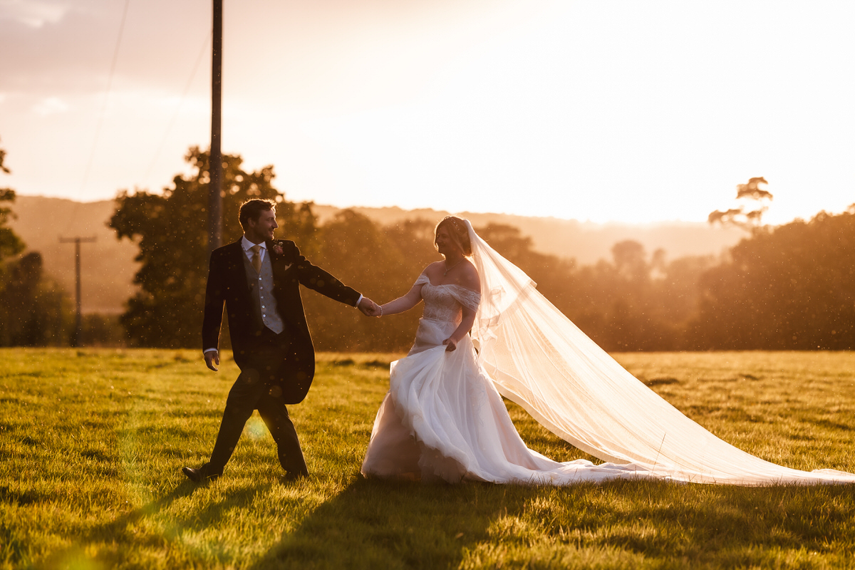 A bride and groom walk hand in hand across a sunlit grassy field at sunset. The bride’s long veil flows behind her, and the scene glows with warm, golden light. Trees and hills are visible in the background.