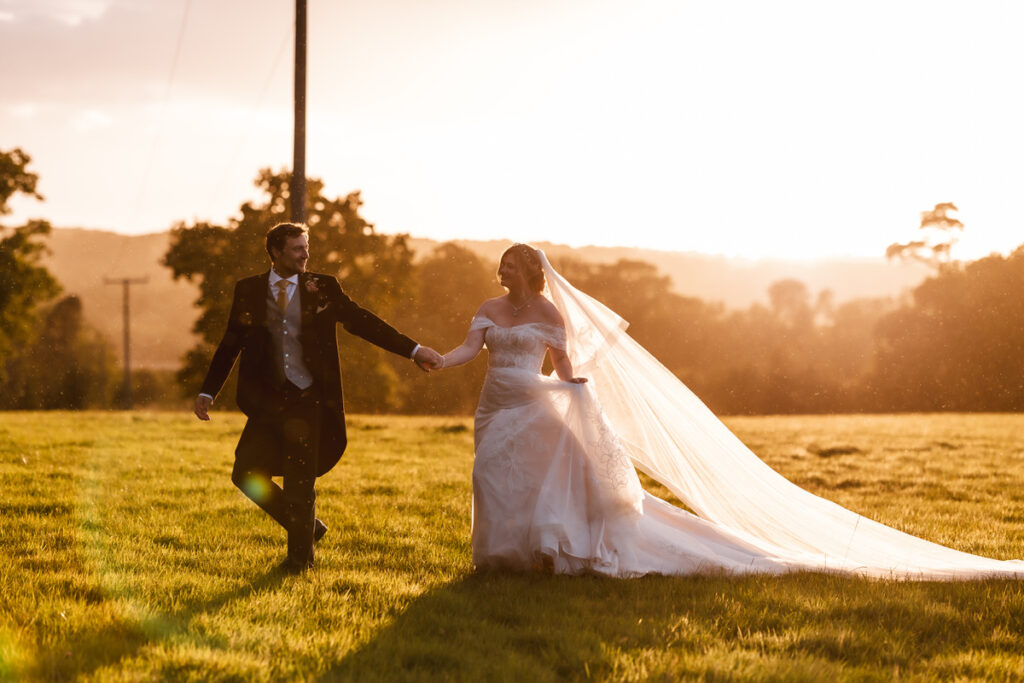 A bride and groom walk hand in hand across a sunlit field at sunset, with the bride’s long veil flowing behind her and golden light casting a warm glow over the scene.