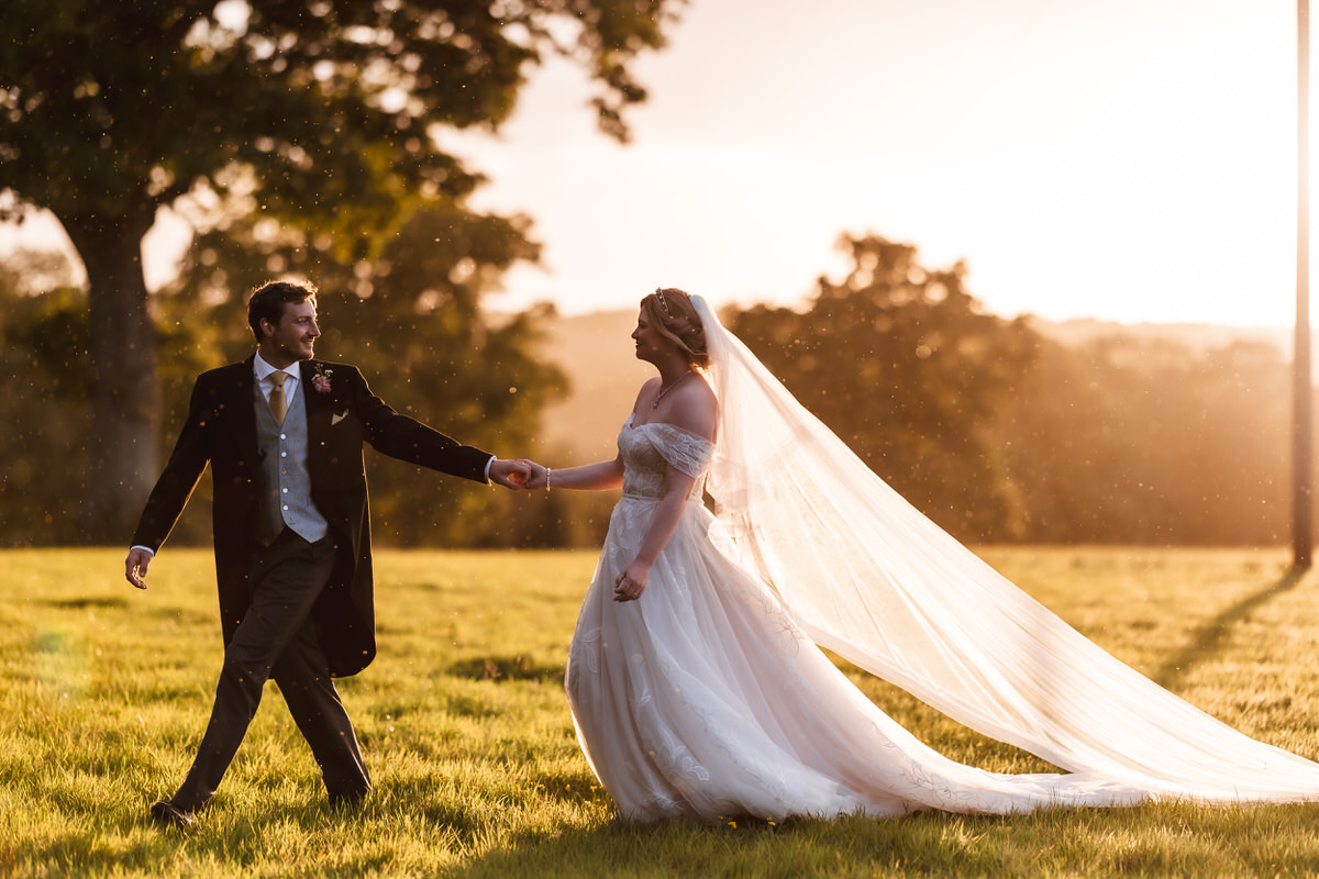 A bride and groom hold hands and smile at each other while walking across a sunlit grassy field. The bride’s long veil flows behind her, and sunlight streams through the trees in the background.