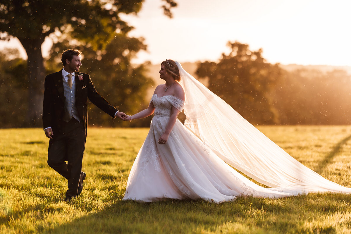 A bride and groom hold hands and smile at each other while walking across a sunlit field. The bride’s long veil flows behind her, and trees are visible in the background under a golden sky.