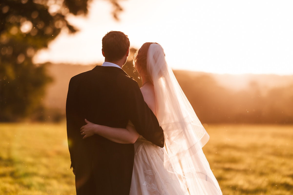 A bride and groom stand arm in arm in a sunlit field at sunset, facing away from the camera. The bride wears a veil and white dress, and the groom wears a dark suit. The scene is warm and peaceful.