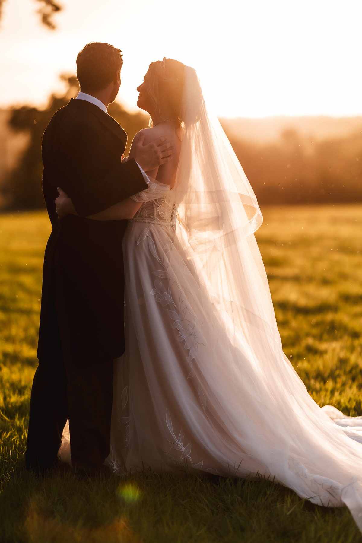 A bride and groom stand arm in arm in a sunlit field at sunset, facing each other and smiling. The bride wears a long white gown and veil, and the groom wears a dark suit.
