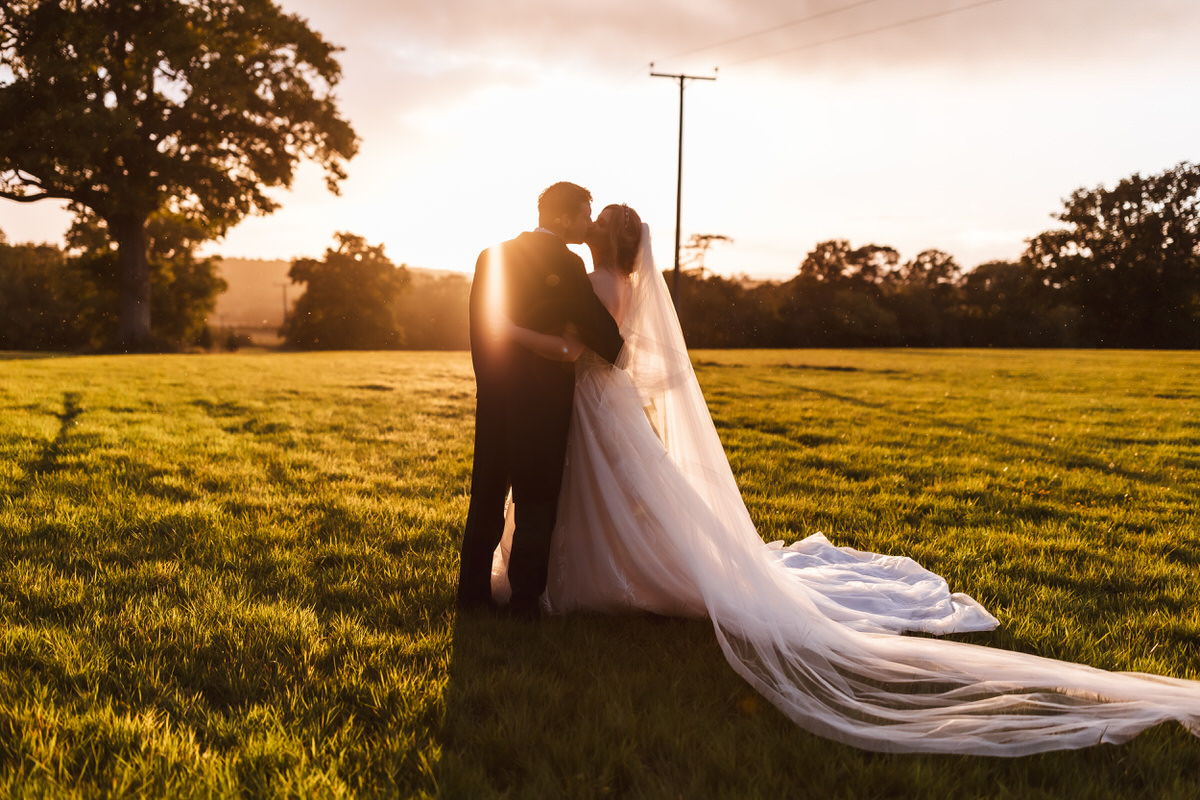 A bride and groom stand close together in a sunlit field at sunset, the bride’s long veil flowing behind her. They are silhouetted against the golden light, surrounded by trees and open grass.
