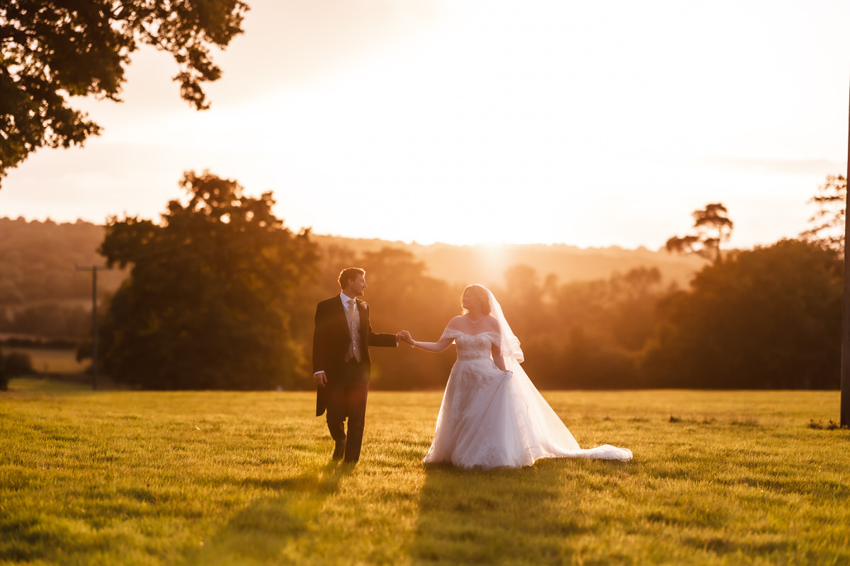 A bride and groom walk hand in hand across a grassy field at sunset, with golden sunlight illuminating their wedding attire and trees in the background.