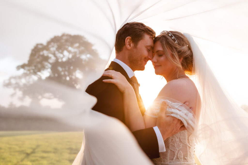 A bride and groom embrace outdoors at sunset, smiling with closed eyes. The bride’s veil flows around them, framing their faces and creating a soft, romantic atmosphere.