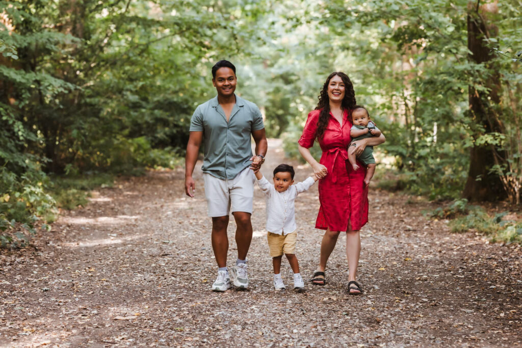 A family of four walks on a forest path. A man and woman hold hands with a young child between them, while the woman carries a baby. They are smiling and surrounded by green trees.