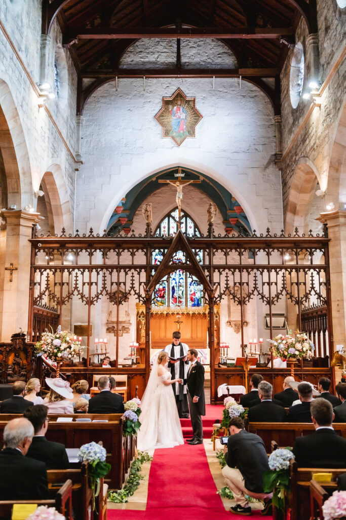 Bride and groom stand together at the altar in a church during their wedding ceremony, with guests seated in pews and ornate decorations surrounding them. The priest stands in front of a stained glass window.