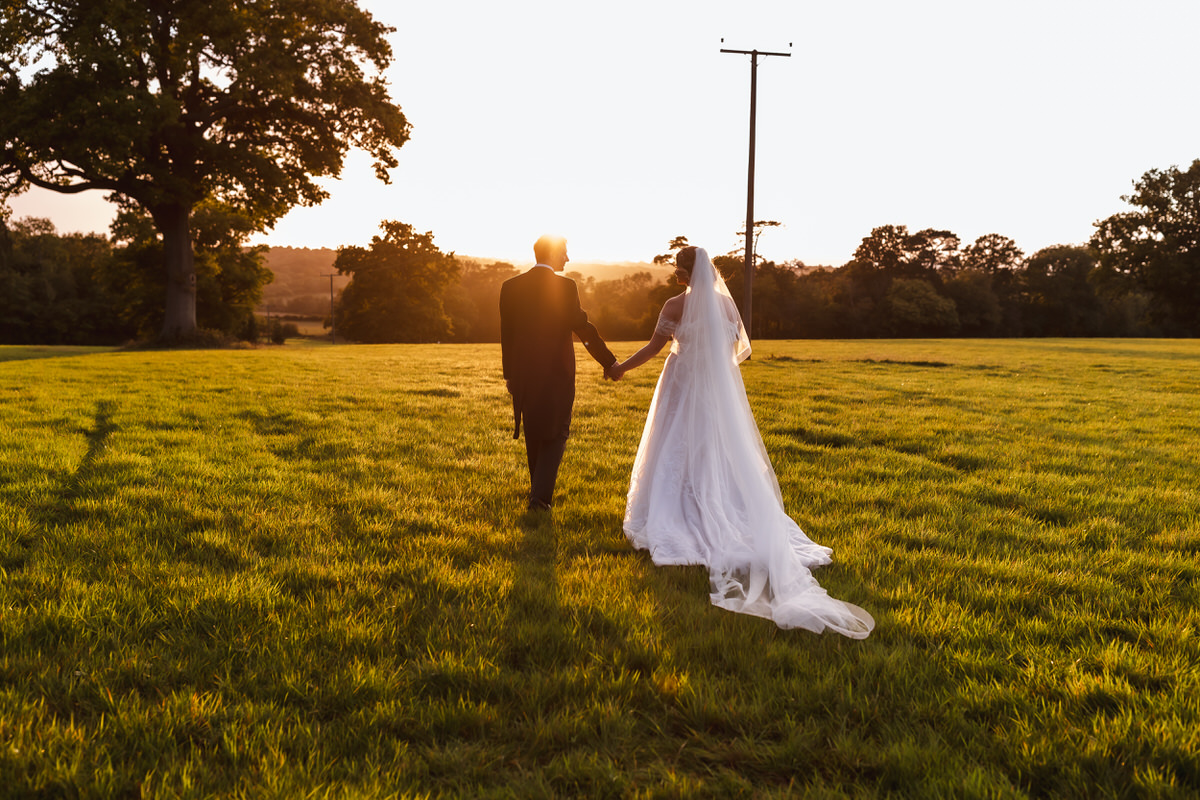 A bride and groom walk hand in hand across a sunlit grassy field at sunset, with trees and power lines in the background. The bride wears a long white dress and veil.