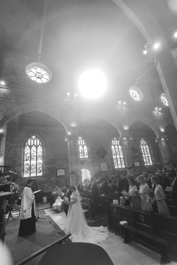 A black-and-white photo of a wedding ceremony inside a church, with the bride and groom standing before an officiant, guests seated in pews, and sunlight streaming through large stained glass windows.