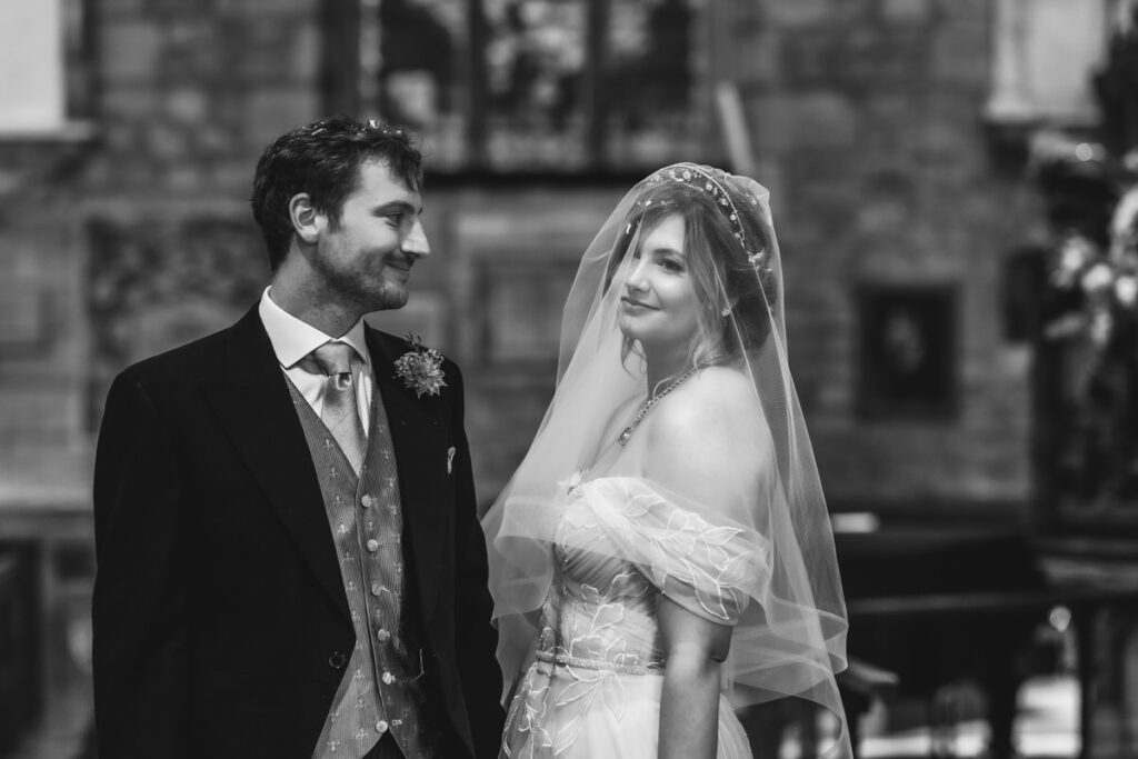 A bride and groom stand together indoors, gazing at each other and smiling. The bride wears a veil and off-the-shoulder dress, while the groom is in a suit with a boutonniere. The background is slightly blurred.