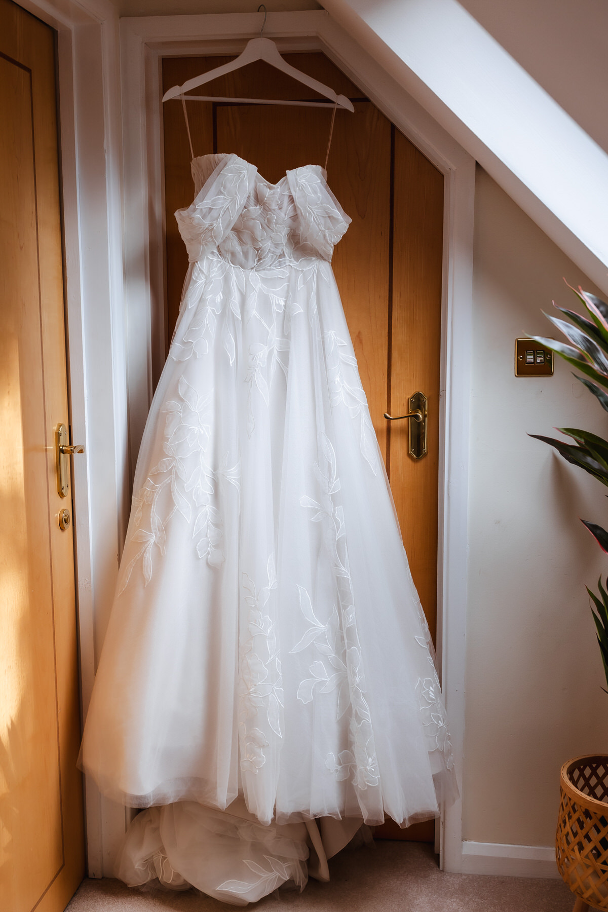 A white wedding dress with floral embroidery hangs on a hanger from a wooden door in a sunlit room, with part of a plant and a woven basket visible on the right.