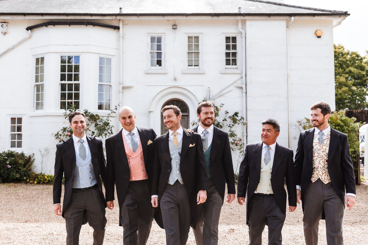 Six men in formal suits with colorful waistcoats walk together outside a large white building, smiling and talking, on a sunny day.