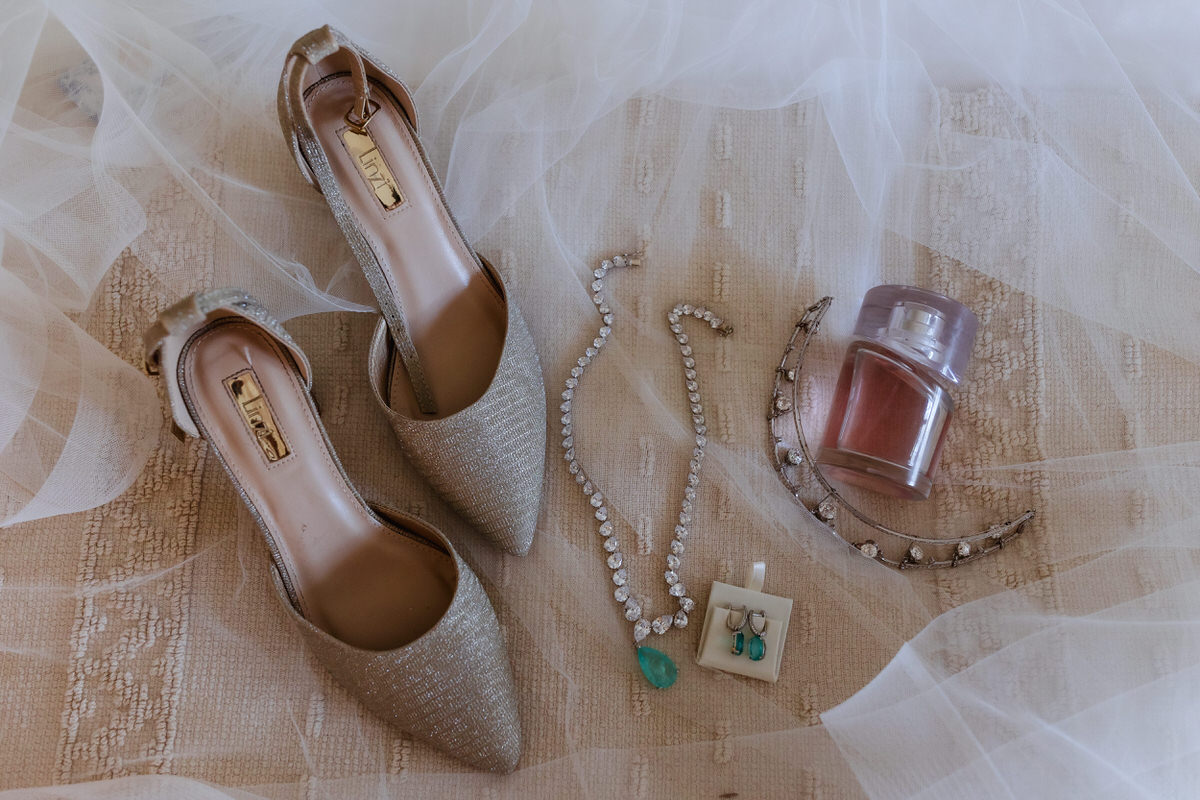 A pair of elegant pointed-toe shoes, a rhinestone necklace with a green pendant, turquoise earrings, a tiara, and a small container of blush are arranged on a soft, textured fabric with sheer veil overlay.