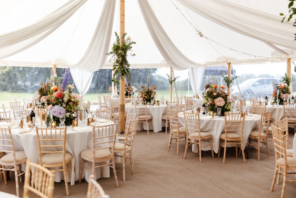 A white tent decorated with draped fabric and greenery shelters round tables with white tablecloths, wooden chairs, and colorful floral centerpieces, creating an elegant event setting.