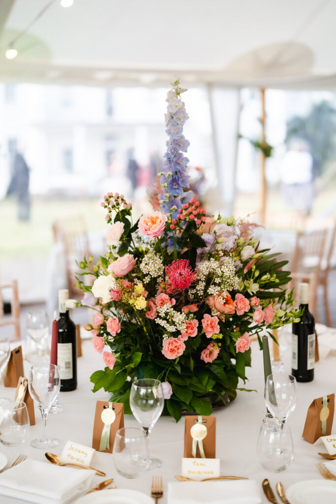A vibrant floral centerpiece with pink, purple, and white flowers sits on a round table set for an event, surrounded by wine bottles, glasses, cutlery, name cards, and small brown gift bags.