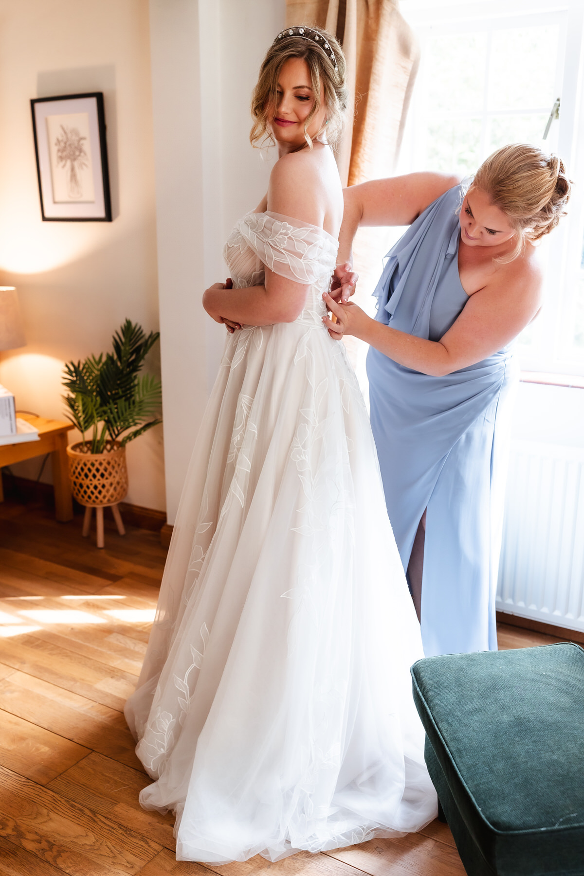 A bride in an off-shoulder white gown smiles as a bridesmaid in a light blue dress helps fasten her dress. They stand in a bright room with wooden floors, a green chair, plants, and framed art on the wall.