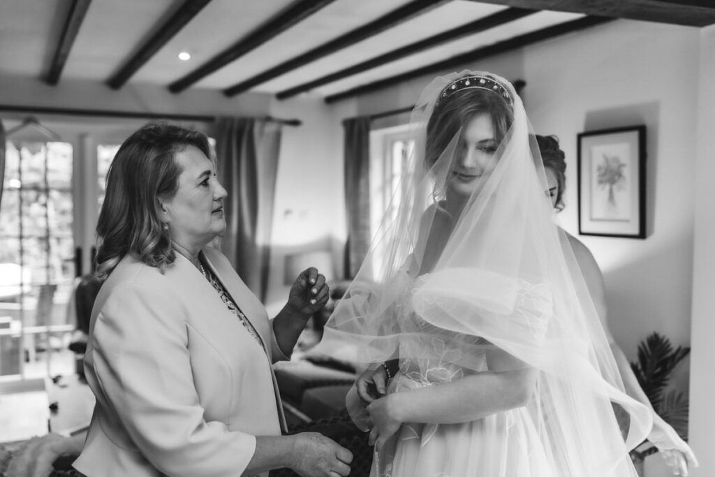 A woman helps a bride adjust her veil indoors. The bride wears a white dress and veil, standing near another person, with natural light coming through windows in the background. The image is in black and white.