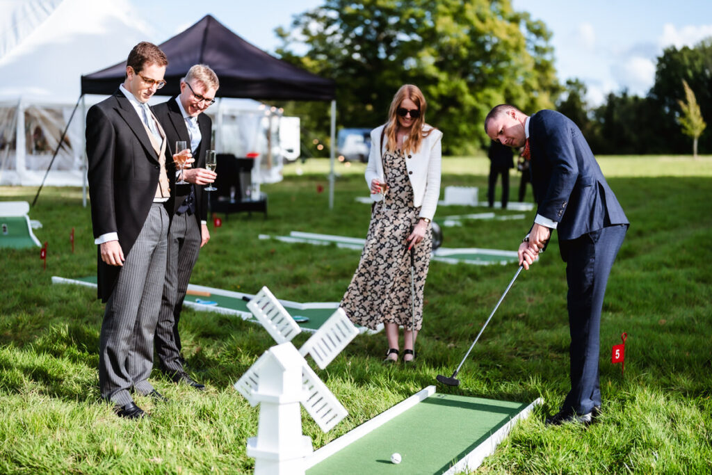 Four people dressed in formal attire play mini golf on a grassy lawn, with one man putting while the others watch, holding drinks. A small windmill obstacle and tents are visible in the background.