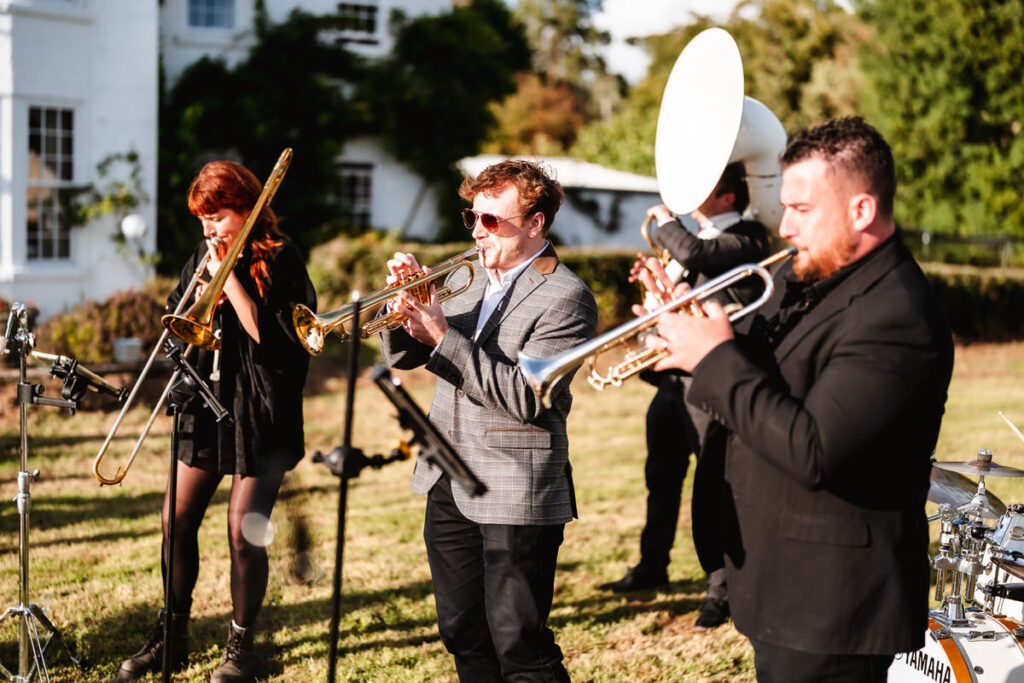 A brass band performs outdoors in sunlight, featuring musicians playing trombone, trumpet, and sousaphone. The group stands on grass, with trees and a white building in the background.