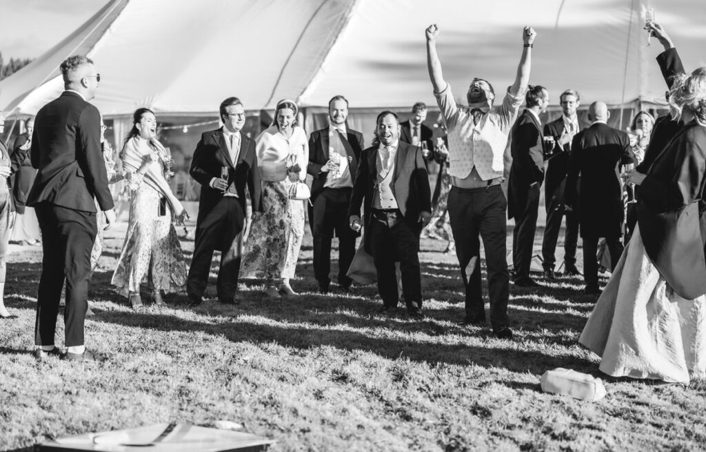 A group of people dressed in formal attire stand on grass near a large tent. One man in the foreground raises his arms in celebration while others smile and watch, suggesting a festive outdoor event. The image is in black and white.