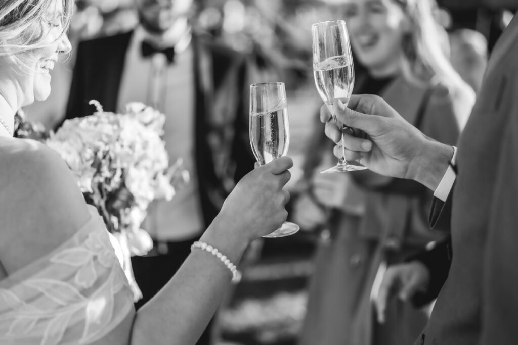 Black and white photo of people dressed formally, holding and clinking champagne glasses in a celebratory toast. One person holds a bouquet of flowers. The mood is festive and joyful.