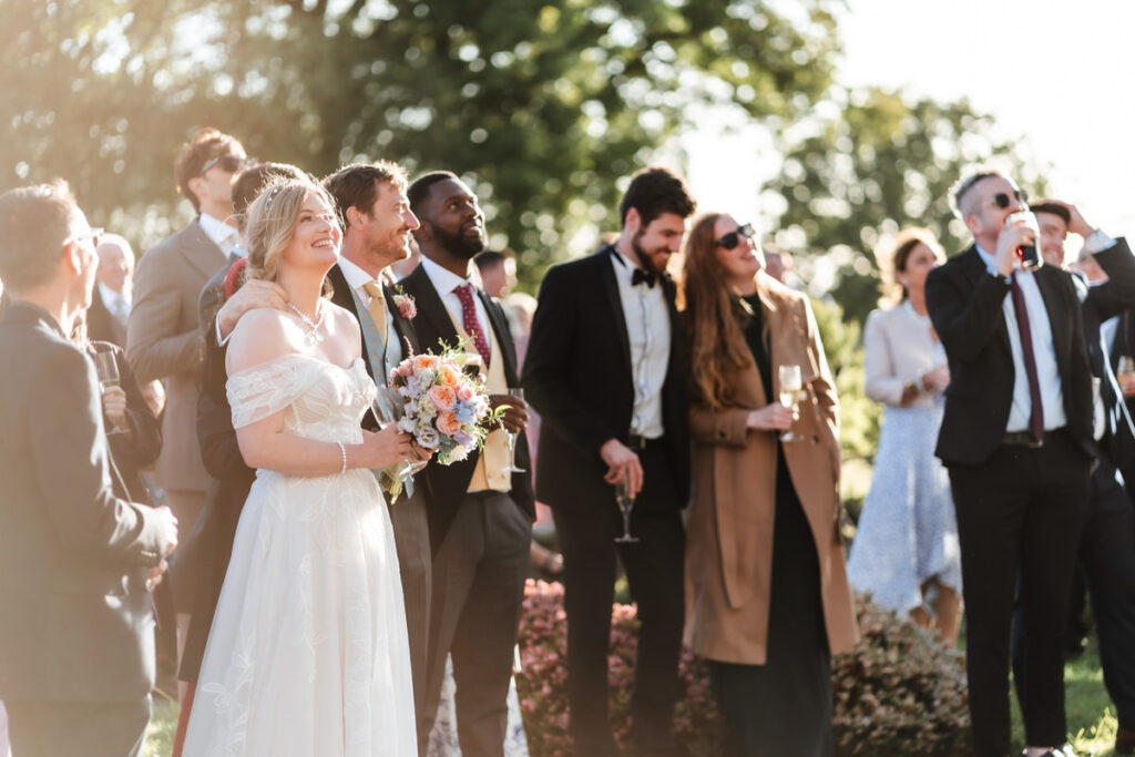 A bride in a white dress stands with a bouquet among well-dressed guests outdoors. People smile, talk, and watch something off camera, enjoying a sunny celebration surrounded by greenery.