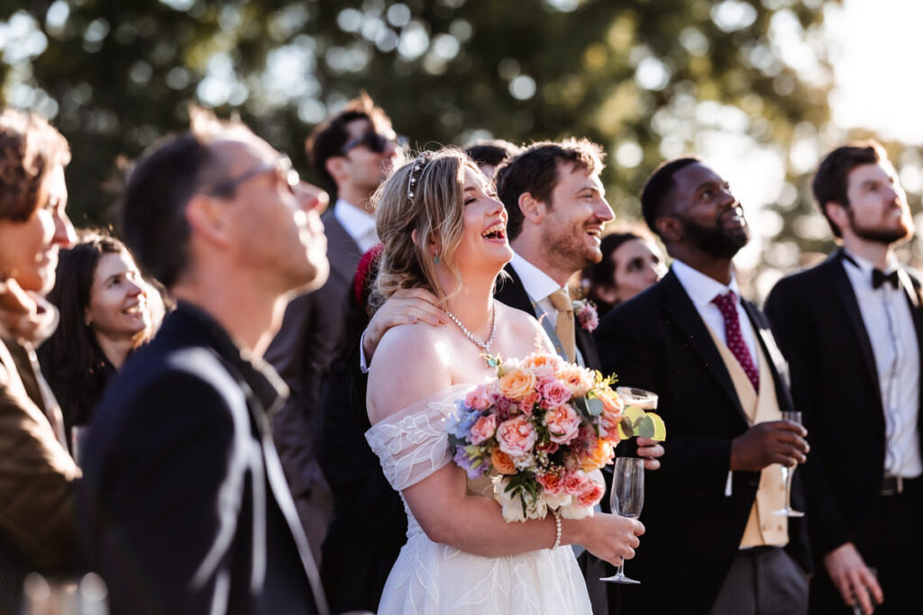 A smiling bride holding a bouquet stands among guests in formal attire at an outdoor wedding, all looking up and appearing joyful. Sunlight filters through trees in the background.