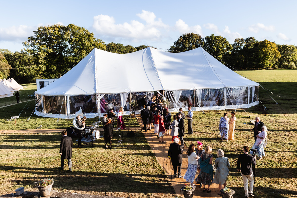 A group of people in formal attire gather outside a large white event tent set up in a grassy field on a sunny day, with trees and blue sky in the background.