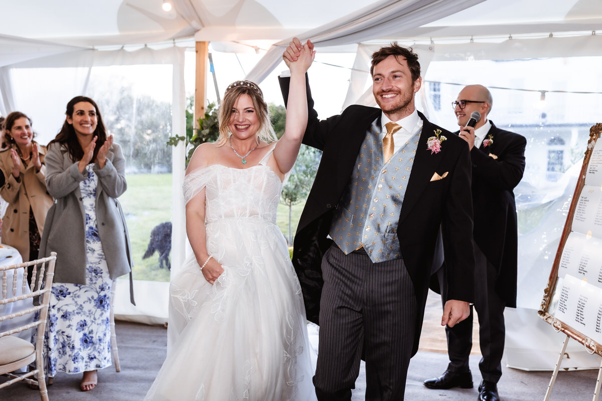 A bride and groom, smiling and holding hands raised in celebration, walk into a tented wedding reception as guests applaud and a man with a microphone stands behind them.