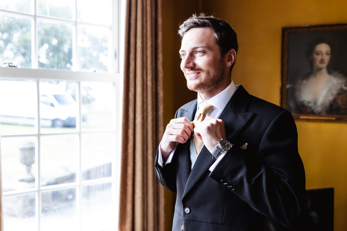 A man in a suit smiles as he adjusts his tie while standing by a bright window. The room has yellow walls, brown curtains, and a framed portrait of a woman on the wall behind him.