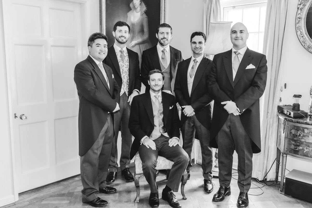 Six men in formal suits pose for a group photo indoors; five stand behind while one sits in a chair. The room has classic decor, including a framed portrait and a mirror on the wall. The image is in black and white.
