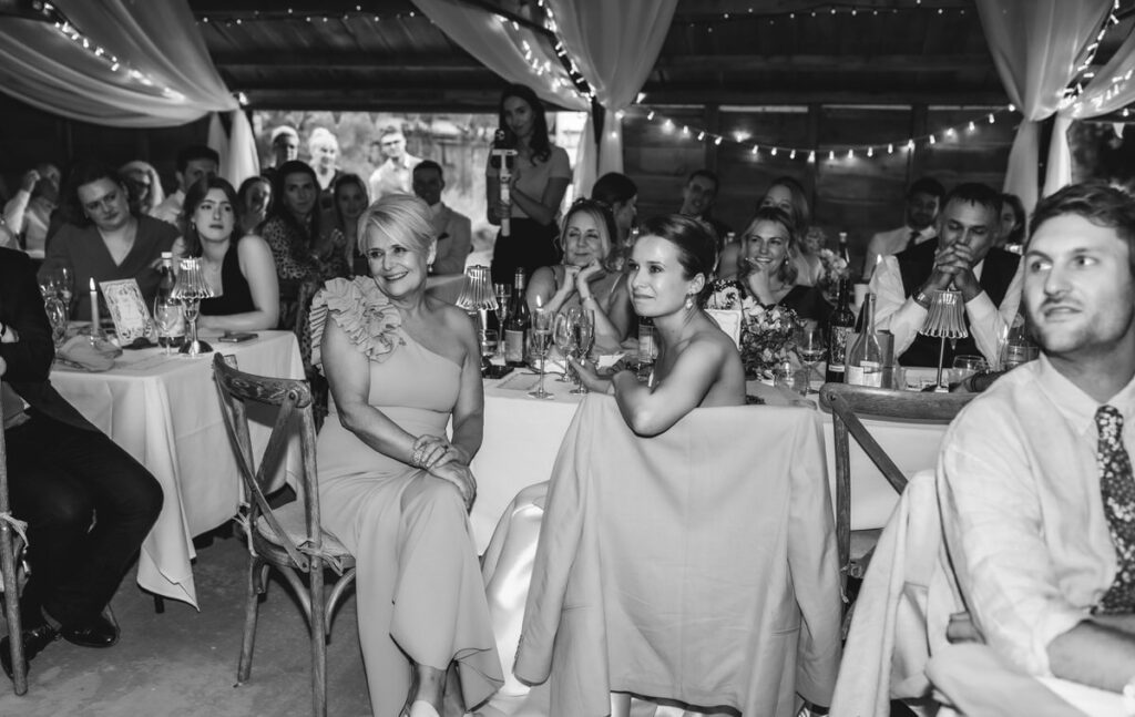 A black and white photo of people seated at banquet tables, dressed formally, attentively watching something. String lights and draped curtains decorate the venue, creating a festive atmosphere.