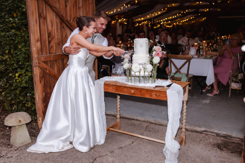 A bride and groom, smiling, cut a white wedding cake together on a wooden table decorated with flowers, while guests watch from decorated tables under string lights in a rustic venue.
