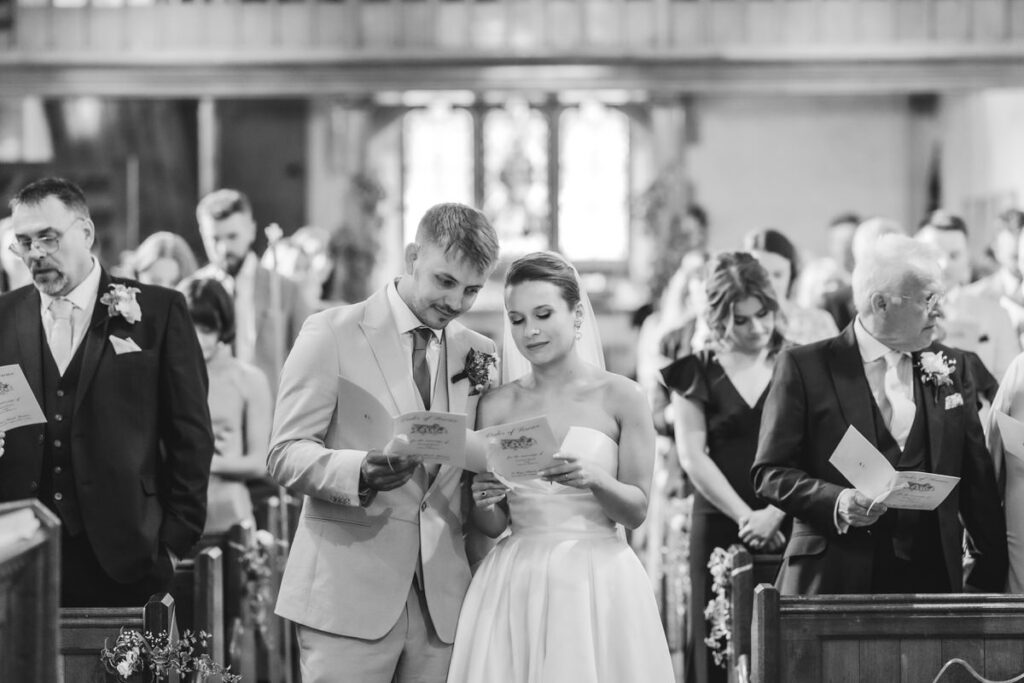 A bride and groom stand together in a church, reading from papers during their wedding ceremony. Guests, dressed formally, are seated and standing behind them. The image is in black and white.