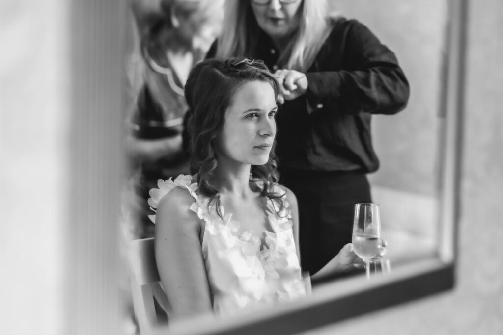 A woman sits in front of a mirror holding a glass, while another person stands behind her styling her hair. The image is in black and white.