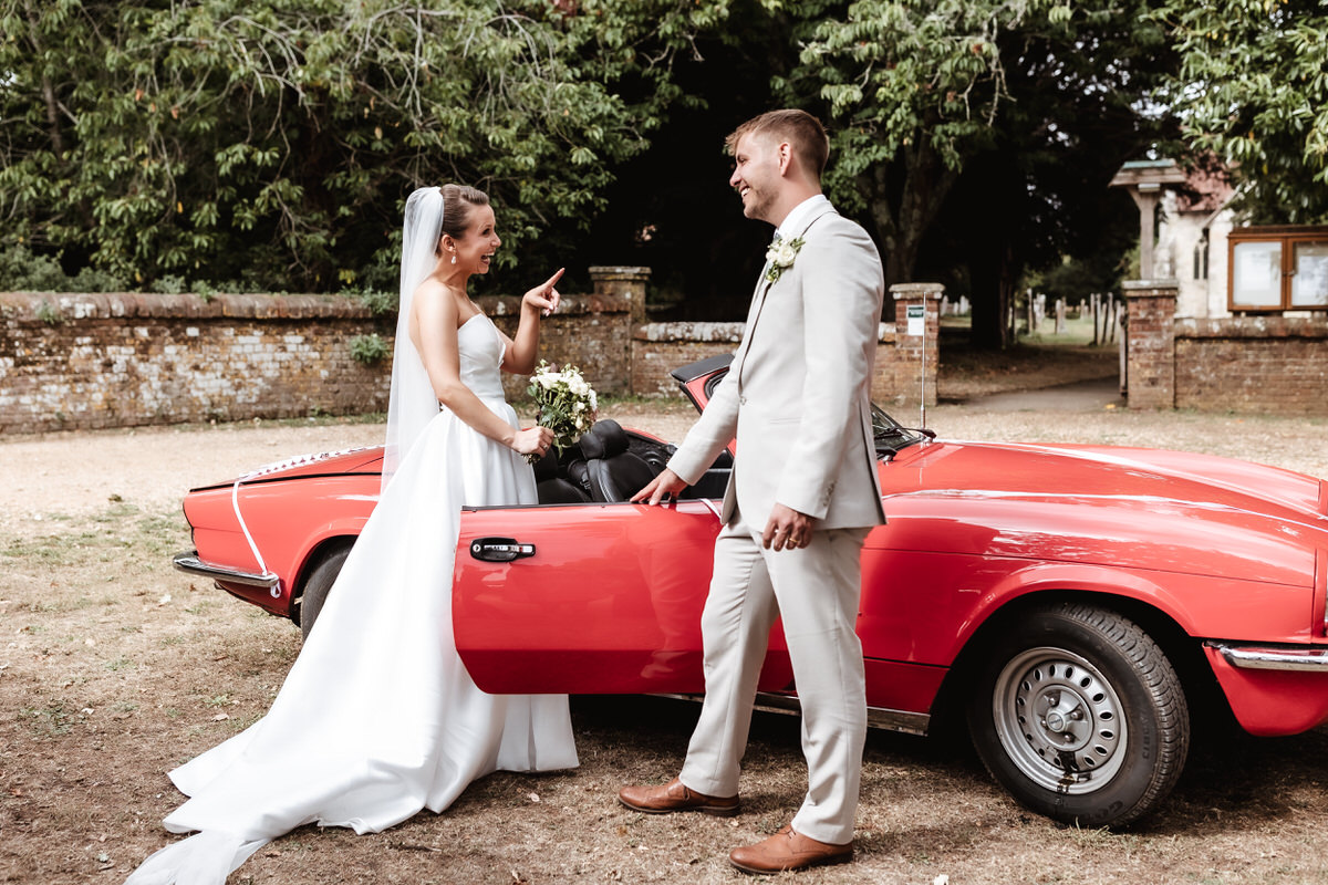 A bride and groom in wedding attire smile and laugh beside a red vintage convertible car in an outdoor setting with trees and a brick wall in the background.