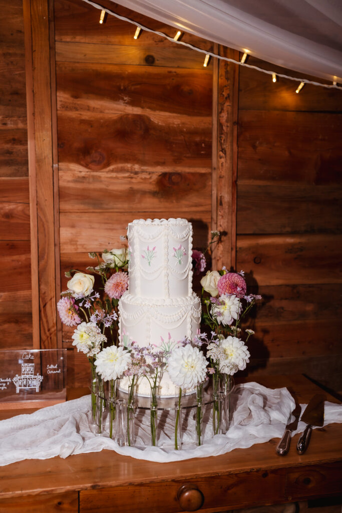 A three-tiered white wedding cake with floral decorations sits on a wooden table, surrounded by fresh white and pink flowers in glass vases, against a wooden wall backdrop. Cake-cutting utensils are nearby.