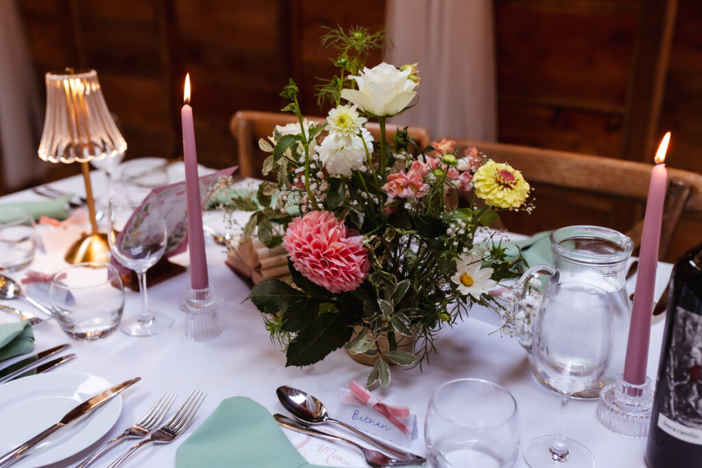 A candlelit table set for a meal, featuring a floral centerpiece with pink, white, and yellow flowers, green napkins, glassware, cutlery, a glass water pitcher, and a small lamp.